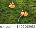 Two yellow oscillating sprinklers water a lush green lawn in a backyard garden. Backyard garden sprinklers irrigate grass on a bright summer day, evoking spring and summer holidays. 140208968