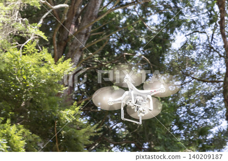 A white drone with rotating propellers flies through the air against a backdrop of green trees. A white drone with rotating propellers flies through the air against a backdrop of green trees. 140209187