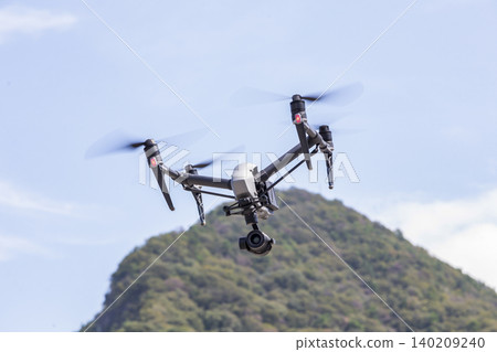 A professional drone flying straight ahead against a backdrop of green mountains. A professional drone flying straight ahead against a backdrop of green mountains. 140209240