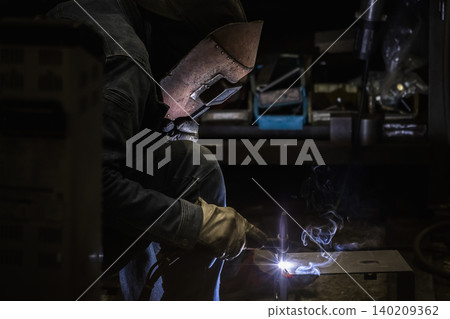 A scene of a craftsman wearing a welding mask welding metal in a darkened factory. A scene of a craftsman wearing a welding mask welding metal in a darkened factory. 140209362