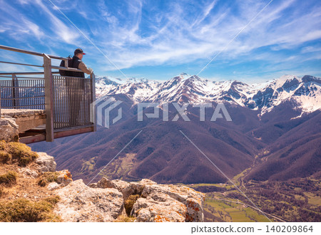 A man tourist stands on a viewpoint above a valley against mountains covered with the snow. A man looks at a beautiful panorama of the mountains. National Park Peaks of Europe (Picos de Europa). A man tourist stands on a viewpoint above a valley against mountains covered with the snow. A man looks at a beautiful panorama of the mountains. National Park Peaks of Europe (Picos de Europa). 140209864