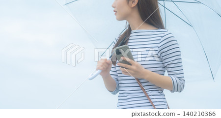 一位女士撐著傘,手裡拿著一支智慧型手機。 一位女士撐著傘,手裡拿著一支智慧型手機。 140210366