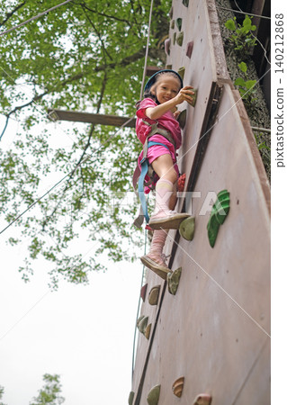 A girl in a protective helmet with a safety rope climbs the wall of a climbing wall in an open-air forest A girl in a protective helmet with a safety rope climbs the wall of a climbing wall in an open-air forest 140212868