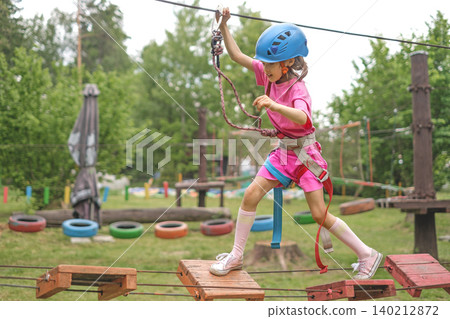 A child steps on the steps of a suspension bridge in a rope entertainment sports town. The girl holds on to the safety rope, moving the carbine 140212872