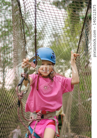The child moves the carbine along the safety rope passing through the grid on the obstacle course. Sports summer entertainment camp for children The child moves the carbine along the safety rope passing through the grid on the obstacle course. Sports summer entertainment camp for children 140212875
