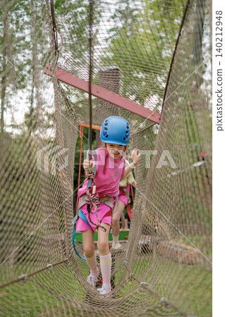 A happy child walks through a tunnel made of mesh. Hanging Rope Obstacle Course in Outdoor Amusement Park A happy child walks through a tunnel made of mesh. Hanging Rope Obstacle Course in Outdoor Amusement Park 140212948