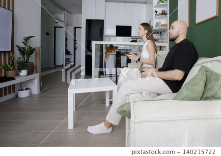 Cute young happy couple playing video games on sofa in modern white and green interior at home Cute young happy couple playing video games on sofa in modern white and green interior at home 140215722