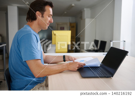 Man working at laptop and reviewing business reports at a bright office desk Man working at laptop and reviewing business reports at a bright office desk 140216192