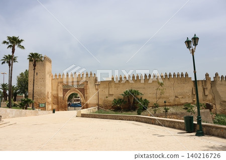 Historic fortified city gate with decorative archway and crenellated walls, framed by palm trees and a curved pedestrian path under an overcast sky, viewed from ground level. Historic fortified city gate with decorative archway and crenellated walls, framed by palm trees and a curved pedestrian path under an overcast sky, viewed from ground level. 140216726