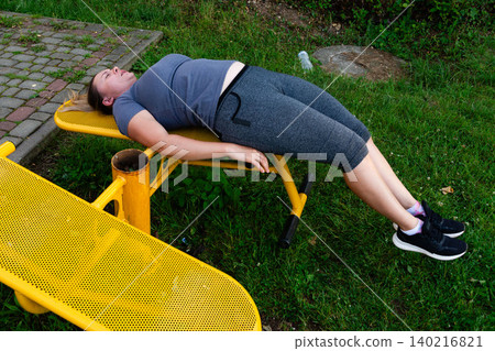 Woman resting on outdoor fitness equipment after a workout. Taking a break in the park. Woman resting on outdoor fitness equipment after a workout. Taking a break in the park. 140216821