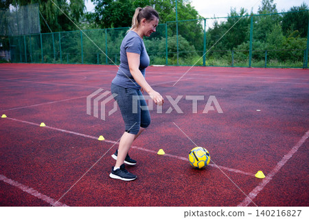 Woman practicing soccer drills on a red court. She dribbles the ball around cones, focusing on agility and ball control. Woman practicing soccer drills on a red court. She dribbles the ball around cones, focusing on agility and ball control. 140216827