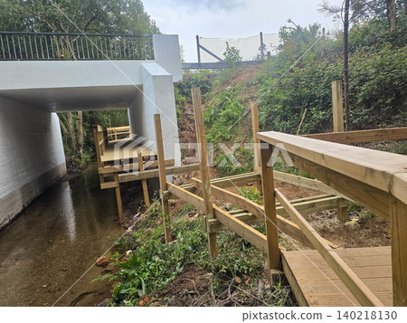 Wooden pedestrian bridge construction near a forest stream with unfinished decking and support posts. Concepts of sustainable infrastructure, outdoor engineering, and eco-friendly development. Wooden pedestrian bridge construction near a forest stream with unfinished decking and support posts. Concepts of sustainable infrastructure, outdoor engineering, and eco-friendly development. 140218130