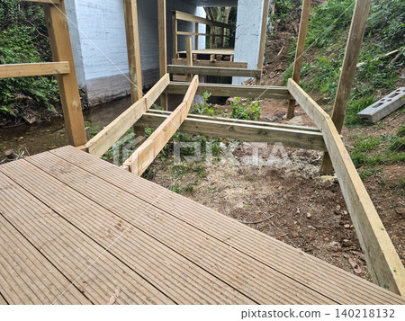 Wooden pedestrian bridge construction near a forest stream with unfinished decking and support posts. Concepts of sustainable infrastructure, outdoor engineering, and eco-friendly development. Wooden pedestrian bridge construction near a forest stream with unfinished decking and support posts. Concepts of sustainable infrastructure, outdoor engineering, and eco-friendly development. 140218132