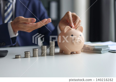 Businessman in a suit putting a coin into a piggy bank with stacks of coins and money on the table. Saving money and financial planning, financial growth, investment, and future planning concept. Businessman in a suit putting a coin into a piggy bank with stacks of coins and money on the table. Saving money and financial planning, financial growth, investment, and future planning concept. 140218282