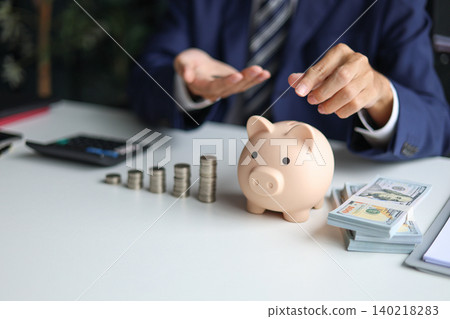 Businessman in a suit putting a coin into a piggy bank with stacks of coins and money on the table. Saving money and financial planning, financial growth, investment, and future planning concept. Businessman in a suit putting a coin into a piggy bank with stacks of coins and money on the table. Saving money and financial planning, financial growth, investment, and future planning concept. 140218283