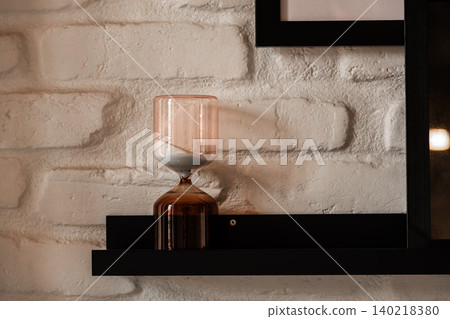 Modern Glass Hourglass on Black Shelf Against White Brick Wall 140218380