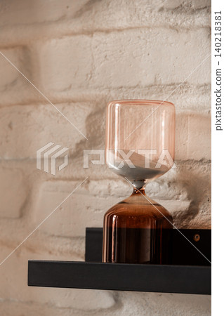 Modern Glass Hourglass on Black Shelf Against White Brick Wall 140218381