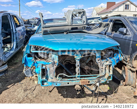 A stripped teal car chassis sits in a scrapyard with a detached plastic dashboard nearby. The scene shows vehicle recycling and the collection of used spare parts during the day. A stripped teal car chassis sits in a scrapyard with a detached plastic dashboard nearby. The scene shows vehicle recycling and the collection of used spare parts during the day. 140218972