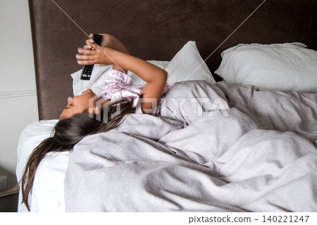 A young girl is laying in bed with a remote in her mouth A young girl is laying in bed with a remote in her mouth 140221247