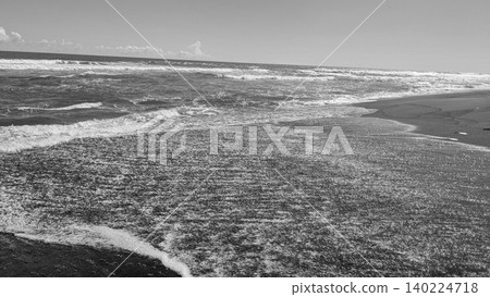 Monochrome high angle view captures glistening ocean water sheet washing over a dark textured sandy beach shore, under a bright daylight sky. Beautiful black white minimalist coastal photography Monochrome high angle view captures glistening ocean water sheet washing over a dark textured sandy beach shore, under a bright daylight sky. Beautiful black white minimalist coastal photography 140224718