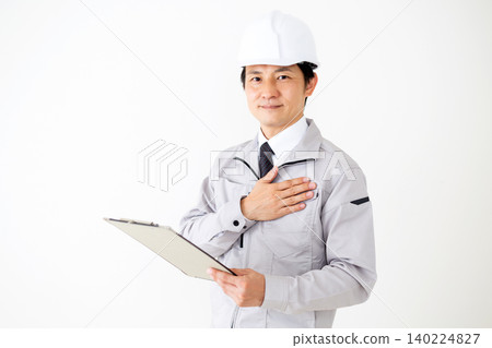 A middle-aged businessman in gray work clothes and a helmet, holding a binder of documents, against a white background. 140224827