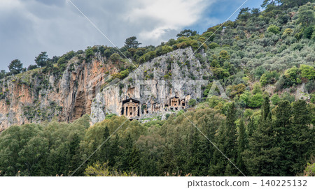 Ancient Lycian rock tombs carved into high cliffs surrounded by lush green trees in Dalyan, Turkey. Historic archaeological landmark and heritage scenery. Ancient Lycian rock tombs carved into high cliffs surrounded by lush green trees in Dalyan, Turkey. Historic archaeological landmark and heritage scenery. 140225132