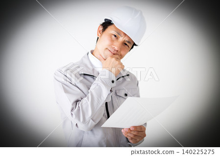 一位身穿灰色工作服、頭戴安全帽的中年商人,在白色背景前若有所思地看著文件。 一位身穿灰色工作服、頭戴安全帽的中年商人,在白色背景前若有所思地看著文件。 140225275