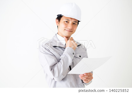一位身穿灰色工作服、頭戴安全帽的中年商人正在白色背景前檢視文件。 一位身穿灰色工作服、頭戴安全帽的中年商人正在白色背景前檢視文件。 140225852