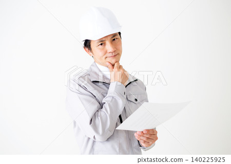 一位身穿灰色工作服、頭戴安全帽的中年商人，在白色背景前若有所思地看著文件。 140225925