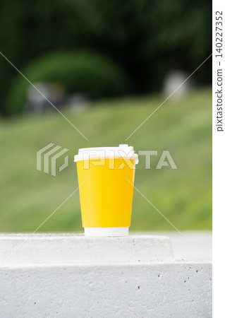 A portrait orientation close-up of a vibrant yellow paper coffee cup with a white lid placed on a concrete ledge outdoors. A beautiful blurred green grass background with plenty of copy space for A portrait orientation close-up of a vibrant yellow paper coffee cup with a white lid placed on a concrete ledge outdoors. A beautiful blurred green grass background with plenty of copy space for 140227352