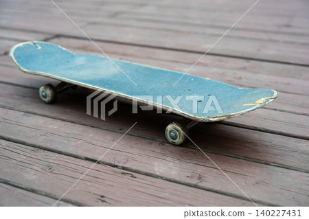 A close-up shot of an old blue skateboard with scratches on a dark wooden board surface. The camera angle is low, showcasing the wheels, trucks, and the texture of the deck. A close-up shot of an old blue skateboard with scratches on a dark wooden board surface. The camera angle is low, showcasing the wheels, trucks, and the texture of the deck. 140227431
