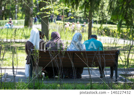 A horizontal medium shot from behind of four unrecognizable people, including women wearing traditional hijab headscarves, relaxing and talking on a wooden bench at an outdoor public park on a bright A horizontal medium shot from behind of four unrecognizable people, including women wearing traditional hijab headscarves, relaxing and talking on a wooden bench at an outdoor public park on a bright 140227432