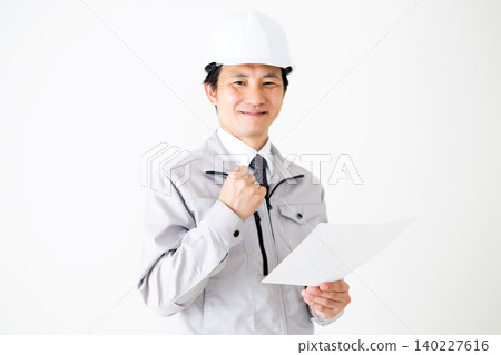 A middle-aged businessman in gray work clothes and a helmet is looking at documents against a white background. 140227616