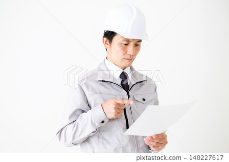 A middle-aged businessman in gray work clothes and a helmet is looking at documents against a white background. A middle-aged businessman in gray work clothes and a helmet is looking at documents against a white background. 140227617