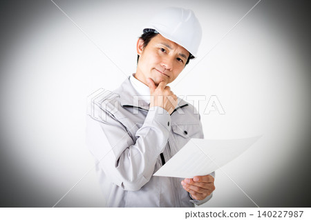 一位身穿灰色工作服、頭戴安全帽的中年商人,在白色背景前若有所思地看著文件。 一位身穿灰色工作服、頭戴安全帽的中年商人,在白色背景前若有所思地看著文件。 140227987