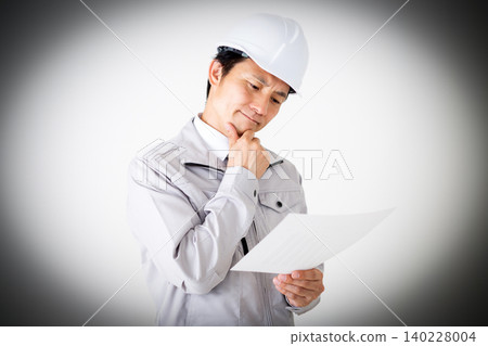 A middle-aged businessman in gray work clothes and a helmet, looking thoughtfully at documents against a white background. A middle-aged businessman in gray work clothes and a helmet, looking thoughtfully at documents against a white background. 140228004