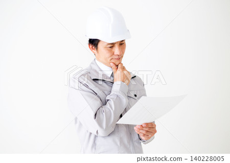 A middle-aged businessman in gray work clothes and a helmet, looking thoughtfully at documents against a white background. A middle-aged businessman in gray work clothes and a helmet, looking thoughtfully at documents against a white background. 140228005