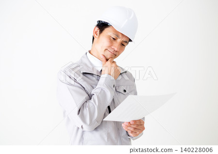 一位身穿灰色工作服、頭戴安全帽的中年商人,在白色背景前若有所思地看著文件。 一位身穿灰色工作服、頭戴安全帽的中年商人,在白色背景前若有所思地看著文件。 140228006