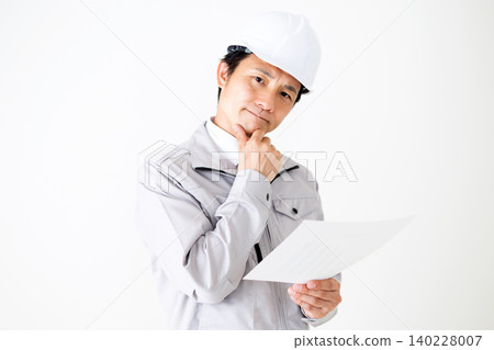 A middle-aged businessman in gray work clothes and a helmet, looking thoughtfully at documents against a white background. A middle-aged businessman in gray work clothes and a helmet, looking thoughtfully at documents against a white background. 140228007