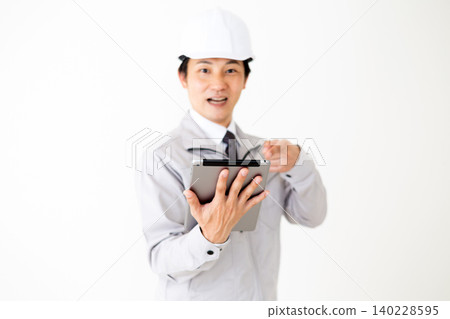 A middle-aged businessman in a gray work uniform and helmet looks surprised while looking at a tablet. (White background) A middle-aged businessman in a gray work uniform and helmet looks surprised while looking at a tablet. (White background) 140228595