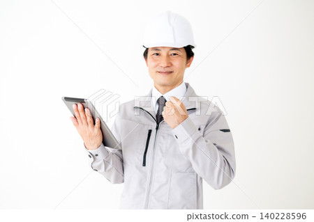 A middle-aged businessman in gray work clothes and a helmet, holding a tablet and making a triumphant gesture, against a white background. A middle-aged businessman in gray work clothes and a helmet, holding a tablet and making a triumphant gesture, against a white background. 140228596