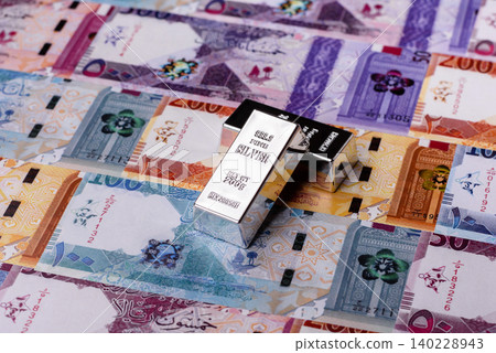 A silver bar, white metal against a backdrop of Qatari money, as a concept. A silver bar A silver bar, white metal against a backdrop of Qatari money, as a concept. A silver bar 140228943