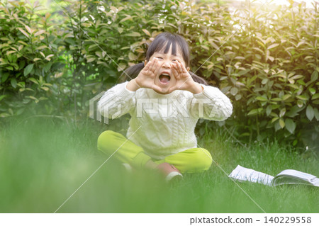 A cute Asian girl sits on the grass outdoors, making a shouting gesture with her hands. A cute Asian girl sits on the grass outdoors, making a shouting gesture with her hands. 140229558