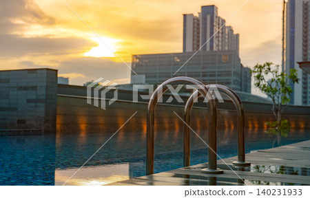Swimming pool at luxury hotel and spa in the morning with golden sunrise sky. Closeup grab bars ladder with clean water and wooden floor at edge of swimming pool. Leisure at hotel poolside concept. Swimming pool at luxury hotel and spa in the morning with golden sunrise sky. Closeup grab bars ladder with clean water and wooden floor at edge of swimming pool. Leisure at hotel poolside concept. 140231933