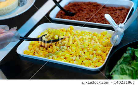 Selective focus on sweet corn in white bowl at salad bar buffet of restaurant. Hand wear plastic gloves holding black spoon scoop yellow sweet corn. Healthy eating. Healthy food. Vegetarian food. Selective focus on sweet corn in white bowl at salad bar buffet of restaurant. Hand wear plastic gloves holding black spoon scoop yellow sweet corn. Healthy eating. Healthy food. Vegetarian food. 140231965