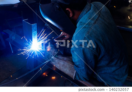 Welder welding metal with argon arc welding machine and has welding sparks. A man wears welding mask and protective gloves. Safety in industrial workplace. Welder working with safety. Steel industry. Welder welding metal with argon arc welding machine and has welding sparks. A man wears welding mask and protective gloves. Safety in industrial workplace. Welder working with safety. Steel industry. 140232057