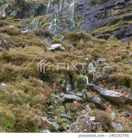 A waterfall cascades down a dark, rocky cliff face overgrown with green moss and vegetation. The lower part of the scene shows a rocky, uneven slope covered in dry brown and green shrubbery. A waterfall cascades down a dark, rocky cliff face overgrown with green moss and vegetation. The lower part of the scene shows a rocky, uneven slope covered in dry brown and green shrubbery. 140233798