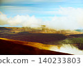 Misty clouds drift over vast brown hills and distant green valleys, with modern wind turbines dotting the horizon on a cool, overcast day. 140233803