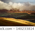 Wind turbines stand tall amidst low-hanging misty clouds above dense green forests and sunlit golden hills. Sunlight illuminates the expansive rural landscape in morning. 140233814