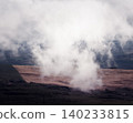 Misty clouds drift low over a vast landscape. A sharp line divides a brown deforested area from a dense dark forest. Wind turbines stand faintly on distant hillsides under the overcast sky. 140233815